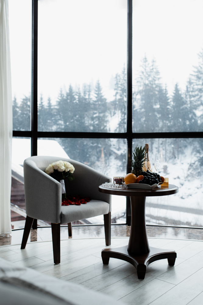 picture of a velvet chair and chair with fruits on top and flower near a window with view of snow and trees